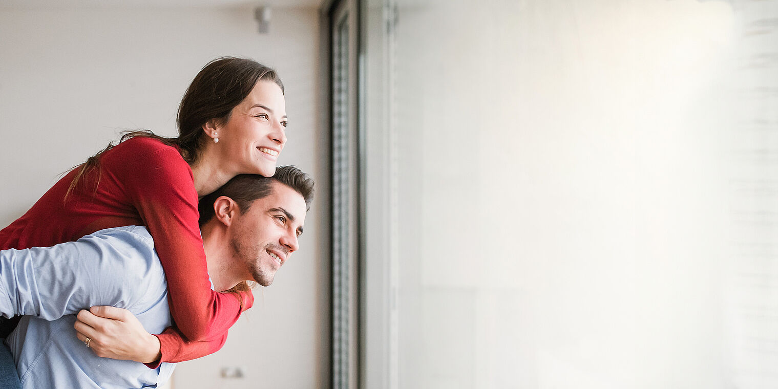 "Happy couple standing by a large window, with the woman embracing the man from behind, both smiling and looking outside