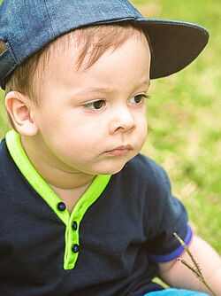 Close-up of a young toddler wearing a cap and a navy shirt with neon green accents, sitting on the grass and looking thoughtful